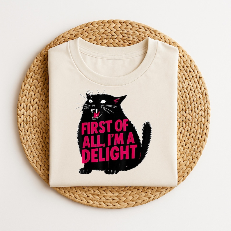 May include: A cream-coloured crew neck t-shirt with a black cat graphic and the pink text "FIRST OF ALL, I'M A DELIGHT". The cat has an open mouth and sharp teeth. The shirt is displayed on a woven, circular placemat, similar to a trivet.