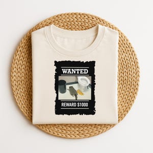 May include: A cream-coloured t-shirt featuring a "Wanted" poster graphic with a raccoon in a bathroom scene. The poster reads "WANTED" and "REWARD £1000". The shirt is presented on a woven, circular placemat.