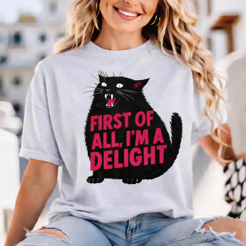 May include: A pale grey t-shirt with a black cat graphic and the pink text "FIRST OF ALL, I'M A DELIGHT". The cat is shown with its mouth open, revealing teeth.