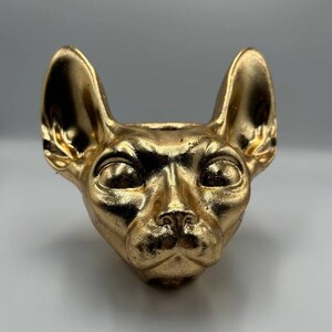 May include: A gold-painted ceramic planter shaped like a Sphynx cat head. The planter has a smooth, glossy finish and realistic details, including the cat's ears, eyes, and nose.