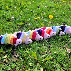 Peut inclure: Plusieurs jouets chenilles crochetés de différentes couleurs, posés sur de l'herbe verte. Chaque chenille présente un schéma de couleurs différent, notamment arc-en-ciel et pastel. Les jouets ont de petits yeux noirs et sont disposés en ligne.