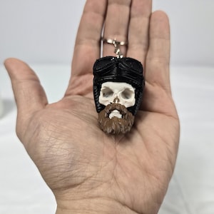 May include: A novelty keychain featuring a skull wearing a black aviator hat and a brown beard. The skull is white with black detailing, and the keychain is attached to a silver ring. The design is detailed and unusual.
