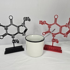 May include: A white enamel mug with a black rim and a silver spoon is centered between two molecular structure models. One model is black, the other red, both labeled "CAFFEINE". The models are on stands.