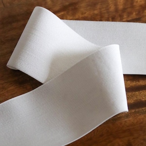 May include: Close-up of white elastic band with a ribbed texture, partially rolled. The elastic is on a wooden surface. The image is well-lit, highlighting the texture and color of the fabric. The elastic is likely used for sewing or crafting projects.