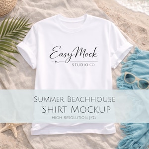 May include: White t-shirt with rolled-up sleeves, featuring the text "Easy Mock Studio Co" in black script. The shirt is on a sandy surface with beach-themed accessories, including sunglasses and a blue towel. The text "Summer Beachhouse Shirt Mockup" is also visible.