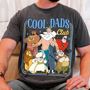 May include: Dark grey t-shirt with a graphic reading "COOL DADS CLUB" in blue and yellow. The graphic features cartoon characters including a lion, a genie, and a superhero.