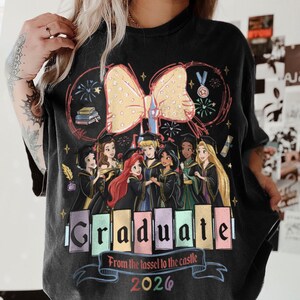 Puede incluir: Camiseta negra con un gr&aacute;fico colorido de princesas de Disney con vestidos de graduaci&oacute;n. La palabra "Graduate" se muestra en letras de bloque coloridas, con el a&ntilde;o 2026 debajo. Un gran lazo amarillo y fuegos artificiales completan el dise&ntilde;o.