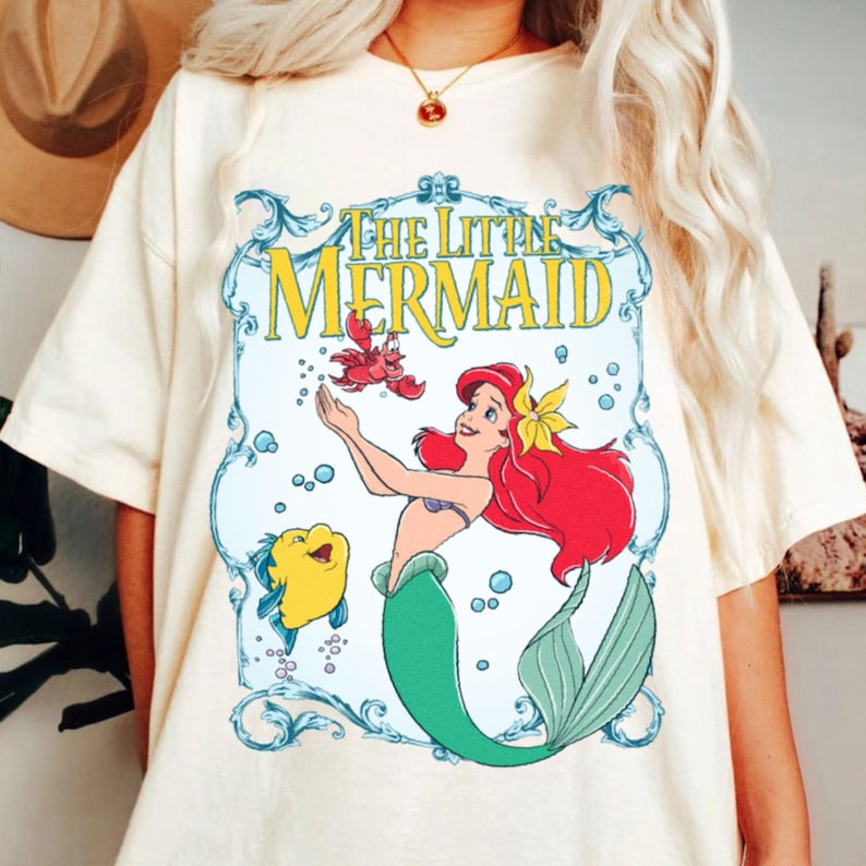 Puede incluir: Camiseta color crema con un gr&aacute;fico de "La Sirenita". El dise&ntilde;o incluye a Ariel con cabello rojo, una cola verde y una flor amarilla, junto con Flounder y Sebasti&aacute;n. Las palabras "The Little Mermaid" se muestran en un marco azul y amarillo.