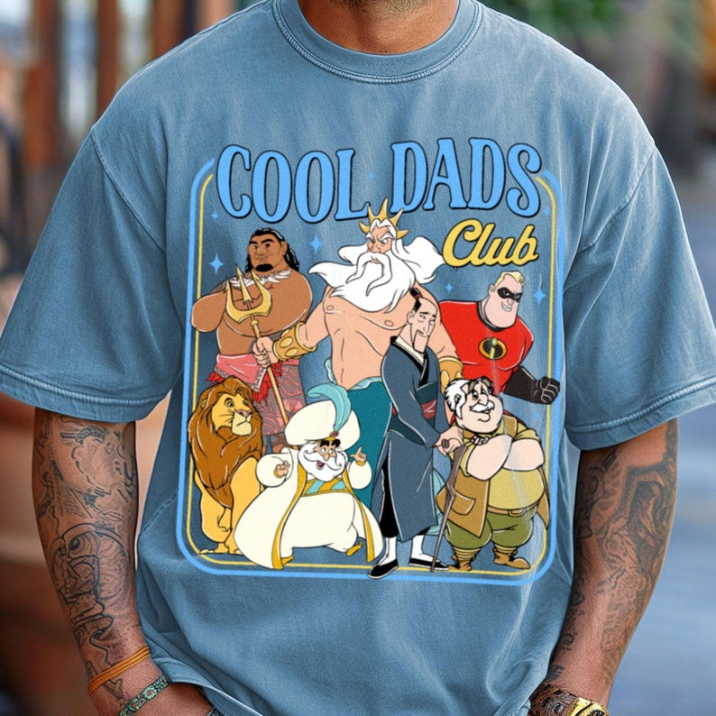 May include: Blue t-shirt with the text "COOL DADS Club" and a graphic featuring cartoon characters. The characters include a lion, a genie, and other figures in a rectangular design with a yellow border.