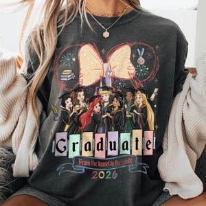 Puede incluir: Sudadera gris oscuro con un gr&aacute;fico de princesas de Disney con vestidos de graduaci&oacute;n. La palabra "Graduate" se muestra en letras de bloque coloridas, con el texto "From the tassel to the castle 2026" debajo. Un gran lazo y fuegos artificiales completan el dise&ntilde;o.
