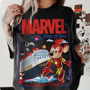 May include: Black t-shirt with a Marvel Day at Sea graphic. The design features Mickey Mouse in an Iron Man suit, a cruise ship, and the text "MARVEL" in red. The shirt has a vintage, distressed look.