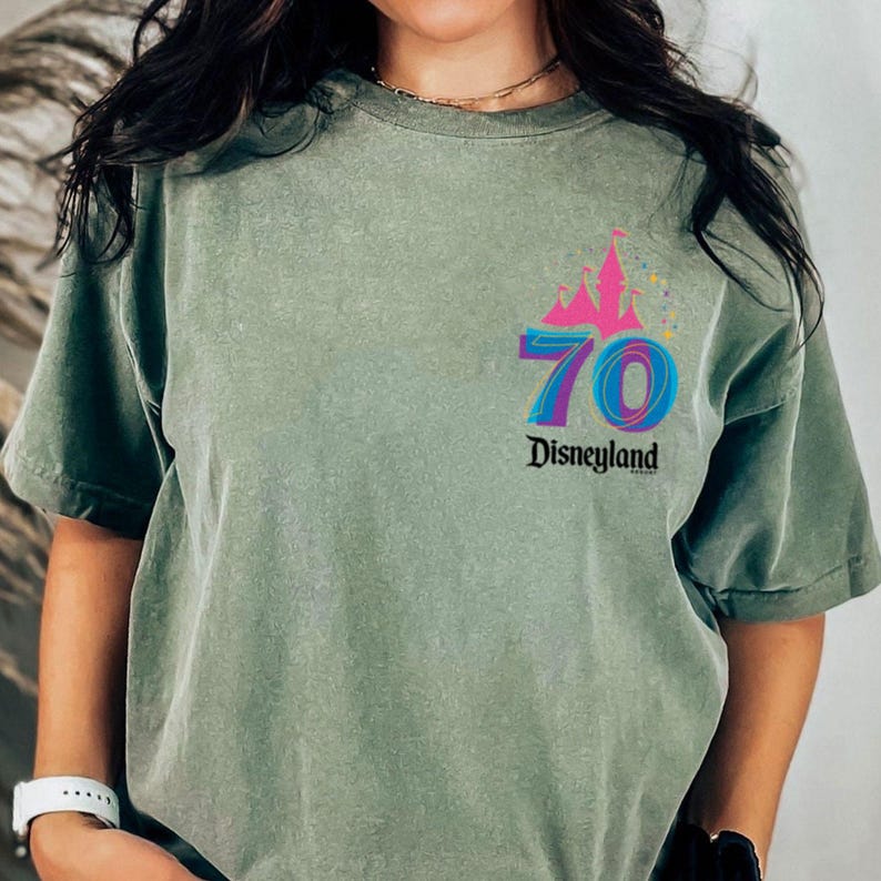 May include: Sage green t-shirt with a colourful graphic celebrating Disneyland's 70th anniversary. The design includes a pink castle, the number 70 in blue and purple, and the word "Disneyland" in black.