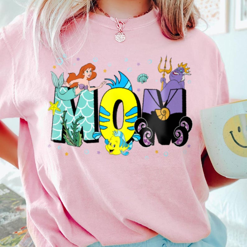 May include: Pink t-shirt with a graphic of the word "MOM" in a whimsical font. The letters are decorated with characters from the Little Mermaid, including Ariel, Flounder, and Ursula.