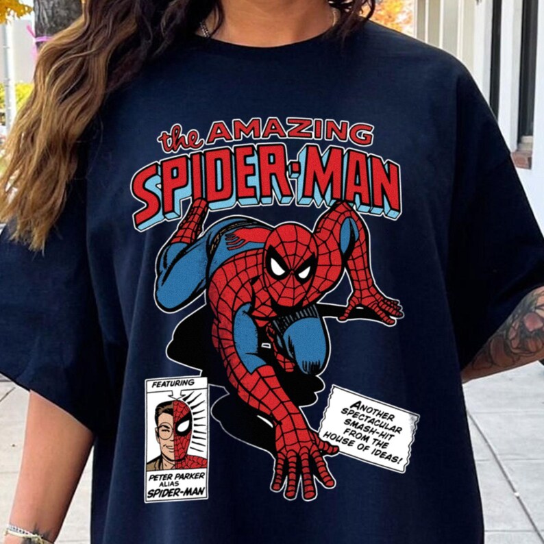 Może przedstawiać: Granatowa koszulka z komiksową ilustracją Spider-Mana. Projekt zawiera tekst "the AMAZING SPIDER-MAN" w kolorze czerwonym i niebieskim oraz dodatkowe dymki. Superbohater jest przedstawiony w swoim klasycznym czerwono-niebieskim kostiumie.