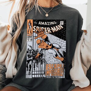 May include: Dark gray sweatshirt featuring a vintage comic book cover of The Amazing Spider-Man. The comic art shows Spider-Man in action with the words "ESCAPE IMPOSSIBLE!" and "The Amazing Spider-Man" printed on the front.