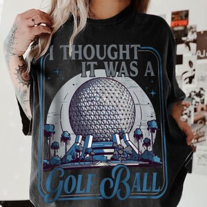 May include: Black t-shirt with a graphic design featuring a geodesic sphere, trees, and the text "I thought it was a Golf Ball" in blue and white. The design is framed in a blue border.