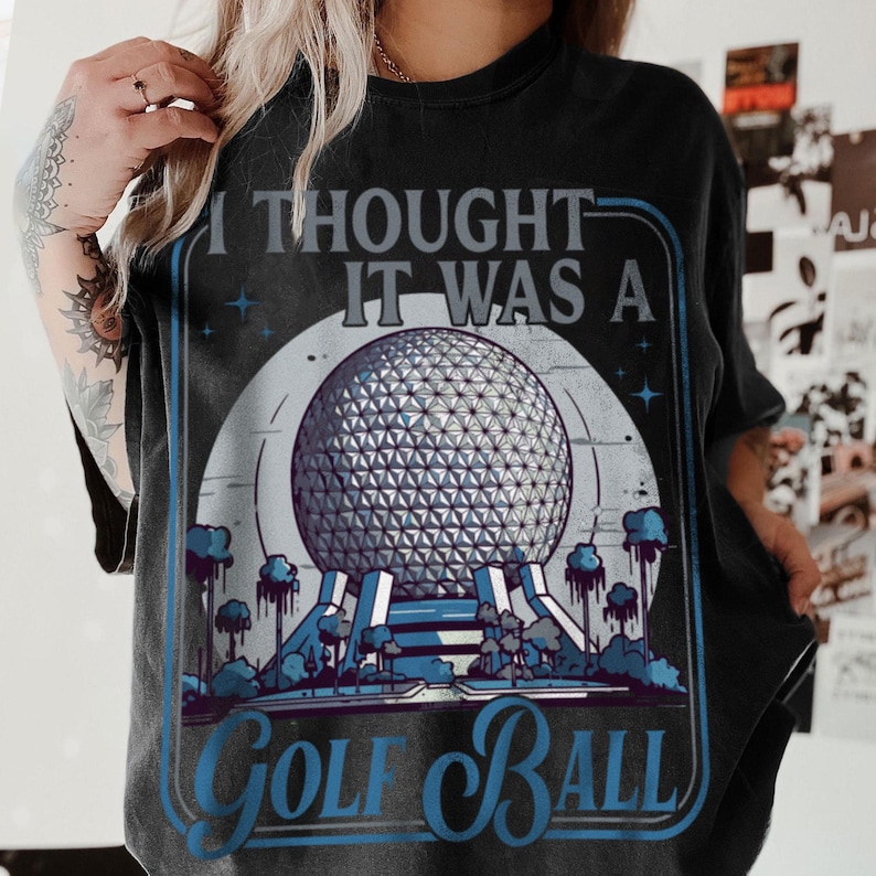 May include: Black t-shirt with a graphic design featuring a geodesic sphere, trees, and the text "I thought it was a Golf Ball" in blue and white. The design is framed in a blue border.