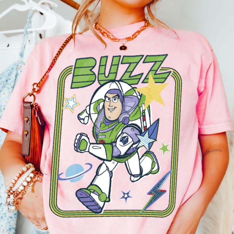 May include: A light pink short-sleeve t-shirt featuring a graphic print of the character Buzz Lightyear in a space suit. The design includes the text BUZZ in green, surrounded by yellow stars, a blue planet, and a blue lightning bolt graphic.