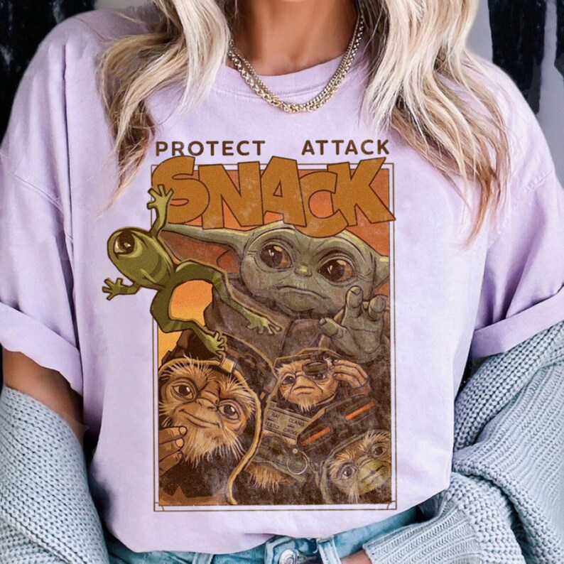 May include: Lilac-colored t-shirt featuring a graphic of a green alien, a baby alien, and other creatures. The text "PROTECT ATTACK SNACK" is printed in brown above the image. The shirt has short sleeves.