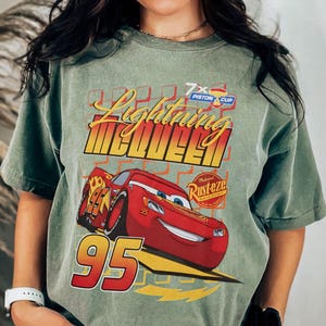 May include: Sage green t-shirt featuring a graphic of Lightning McQueen from the movie Cars. The design includes the text "Lightning McQueen" in yellow and red, the number "95", and the Rust-eze logo.