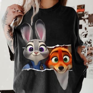 May include: Black t-shirt featuring a graphic of Judy Hopps and Nick Wilde from Zootopia. Judy, a gray rabbit, wears a blue uniform. Nick, an orange fox, has green eyes. The design appears torn, revealing the characters.