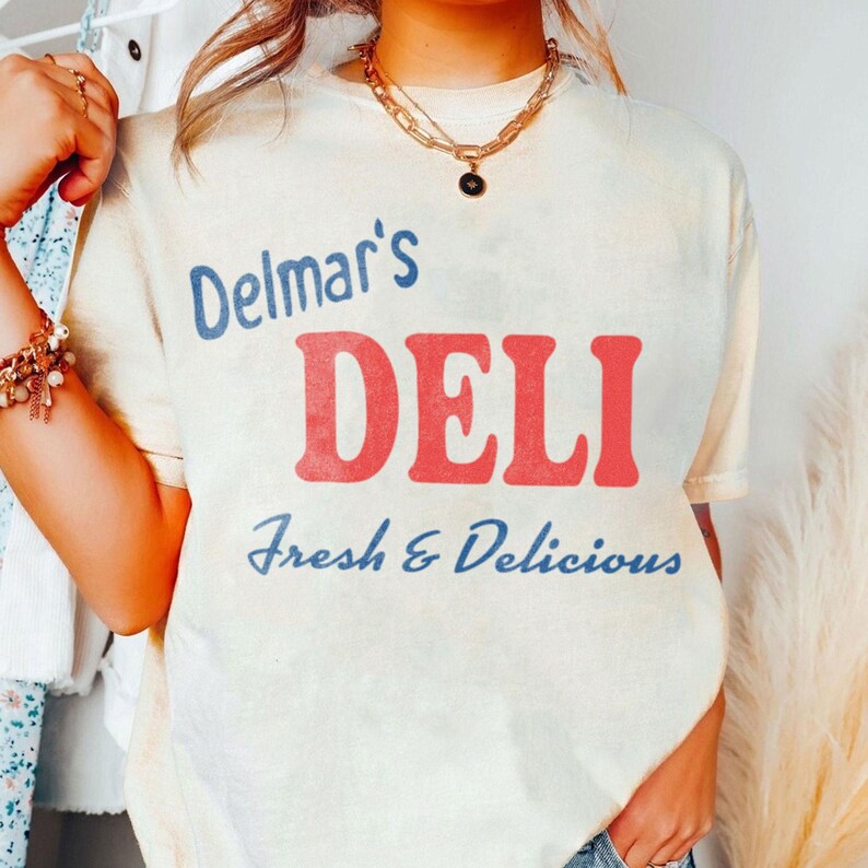 May include: A cream-colored short-sleeve t-shirt featuring the blue text Delmar's, the large red text DELI, and the blue text Fresh & Delicious printed on the front. The shirt has a crew neckline and a relaxed fit, showcasing a casual style.