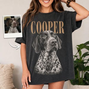 May include: A charcoal gray short-sleeve t-shirt featuring a large graphic of a German Shorthaired Pointer dog with the name COOPER printed in tan capital letters above the dog. A small inset photo shows the same dog in a car.