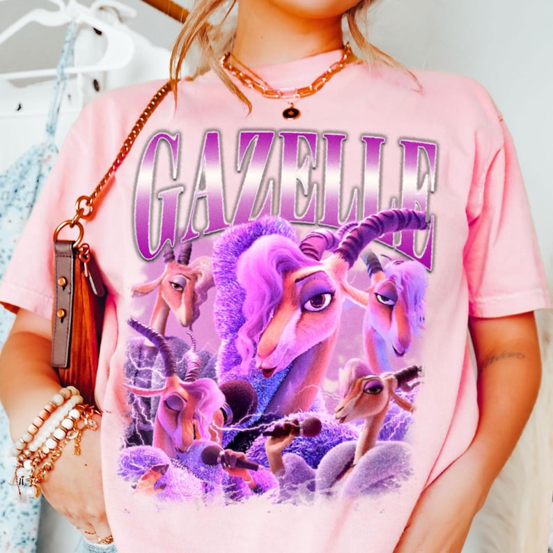 May include: A light pink t-shirt featuring a graphic of a purple gazelle with purple hair and the word "GAZELLE" in a purple and white font. The gazelle is holding a microphone and is surrounded by other gazelles.