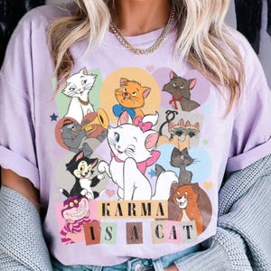 Puede incluir: Una camiseta de color lavanda con un estampado gr&aacute;fico de varios gatos de dibujos animados. El texto "KARMA IS A CAT" est&aacute; impreso en negro en la camiseta.