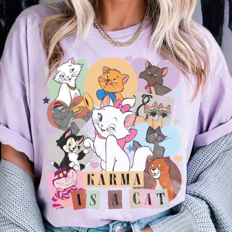 Puede incluir: Una camiseta de color lavanda con un estampado gr&aacute;fico de varios gatos de dibujos animados. El texto "KARMA IS A CAT" est&aacute; impreso en negro en la camiseta.