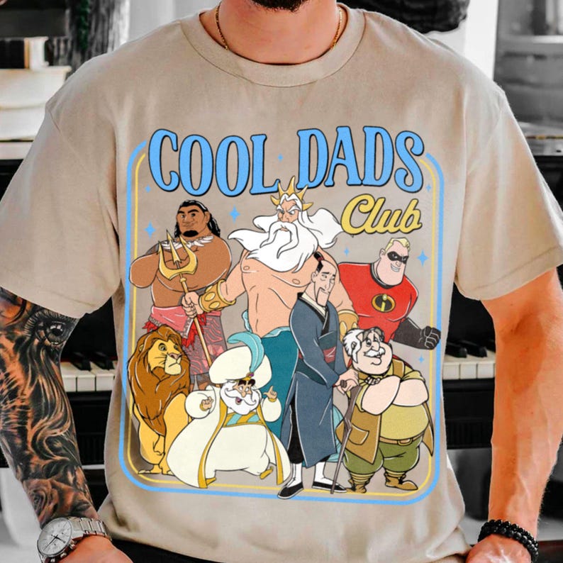 May include: Beige t-shirt featuring a cartoon graphic and the text "COOL DADS Club" in blue. The design includes various animated characters, such as a lion, a man with a trident, and a superhero.