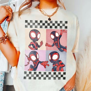 Puede incluir: Camiseta de color crema con un gr&aacute;fico de cuatro paneles de un superh&eacute;roe de dibujos animados con un traje negro y rojo. El dise&ntilde;o incluye un borde a cuadros blanco y negro en la parte superior e inferior. El superh&eacute;roe est&aacute; en diferentes poses de acci&oacute;n.