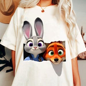 May include: White t-shirt featuring a graphic of Judy Hopps and Nick Wilde from Zootopia. Judy, a gray rabbit with purple eyes, and Nick, an orange fox with green eyes, are depicted peeking through a torn paper effect.