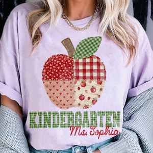 May include: A light purple t-shirt featuring a large apple graphic composed of four distinct patchwork-style sections with red floral, red gingham, heart, and strawberry patterns. Below the apple, the text reads KINDERGARTEN Ms. Sophia.