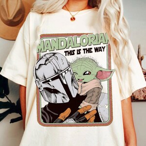 Puede incluir: Camiseta color crema con un gr&aacute;fico de The Mandalorian y Grogu. El dise&ntilde;o incluye el texto "MANDALORIAN THIS IS THE WAY" en verde y blanco. La obra de arte representa a los personajes en un estilo c&oacute;mic.