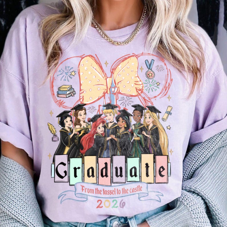 Puede incluir: Camiseta lila claro con un dise&ntilde;o de princesas de dibujos animados con atuendo de graduaci&oacute;n. La palabra "GRADUATE" se muestra en letras de bloque coloridas, con el texto "From the tassel to the castle 2026" debajo.