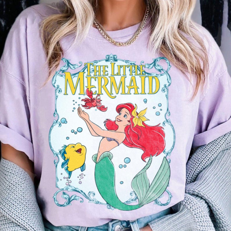 Puede incluir: Una camiseta de color lila claro con un dise&ntilde;o gr&aacute;fico de "La Sirenita". El dise&ntilde;o incluye a Ariel con pelo rojo, una flor amarilla y una cola verde, junto con Flounder y Sebasti&aacute;n. El texto "THE LITTLE MERMAID" es de color amarillo.