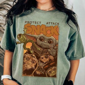 May include: Sage green t-shirt featuring a graphic of Grogu, a frog, and other creatures with the words "PROTECT ATTACK SNACK" in orange and brown. The design is centered on the front of the shirt.