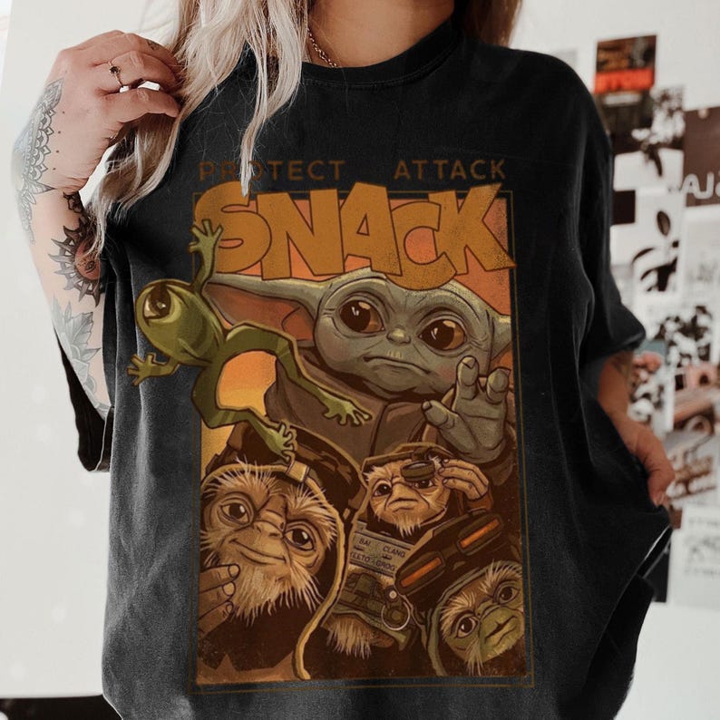May include: Black t-shirt featuring a graphic of a green frog, a character resembling Baby Yoda, and other creatures. The word "SNACK" is prominently displayed in orange letters, with the words "PROTECT ATTACK" above it.