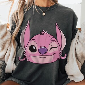 May include: Dark gray sweatshirt featuring a pink cartoon character with large ears, a purple nose, and a winking eye. The character's face is centered on the front of the shirt. The sweatshirt has a crew neck and long sleeves.