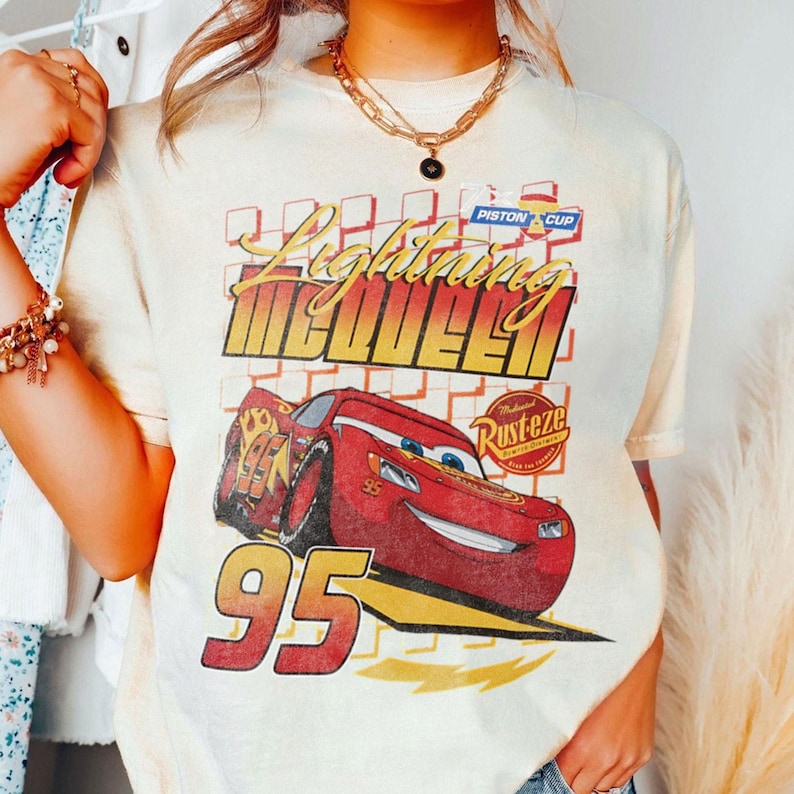 May include: Cream-colored t-shirt featuring a graphic of Lightning McQueen from the movie Cars. The design includes the text "Lightning McQueen" and the number "95" in red and yellow. The shirt has a vintage, retro style.