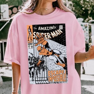 May include: Pink t-shirt featuring a comic book cover of Spider-Man. The graphic shows Spider-Man in action with the text "The Amazing Spider-Man" and "Escape Impossible!" printed on the front. The shirt is a light pink color.