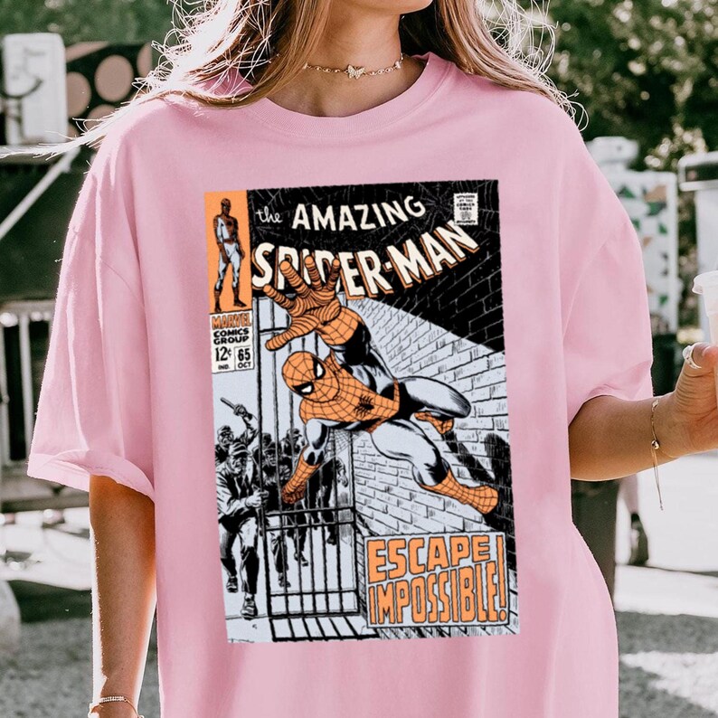 May include: Pink t-shirt featuring a comic book cover of Spider-Man. The graphic shows Spider-Man in action with the text "The Amazing Spider-Man" and "Escape Impossible!" printed on the front. The shirt is a light pink color.