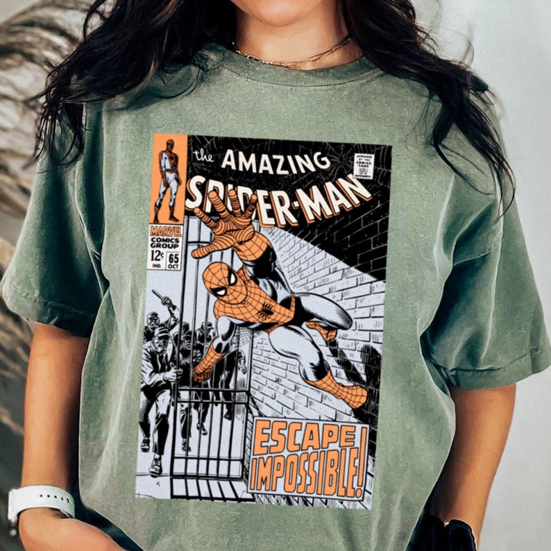 May include: Sage green t-shirt featuring a comic book cover of Spider-Man. The comic book cover has the title "The Amazing Spider-Man" and the words "Escape Impossible!" in orange. The shirt is a comfortable, casual style.