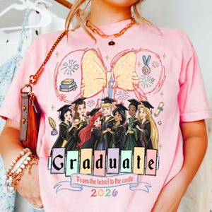 Puede incluir: Camiseta rosa con un dise&ntilde;o de graduaci&oacute;n con princesas de Disney. La palabra "Graduate" se muestra en una fuente colorida, con el texto "From the tassel to the castle 2026" debajo. Un gran lazo amarillo est&aacute; en la parte superior.
