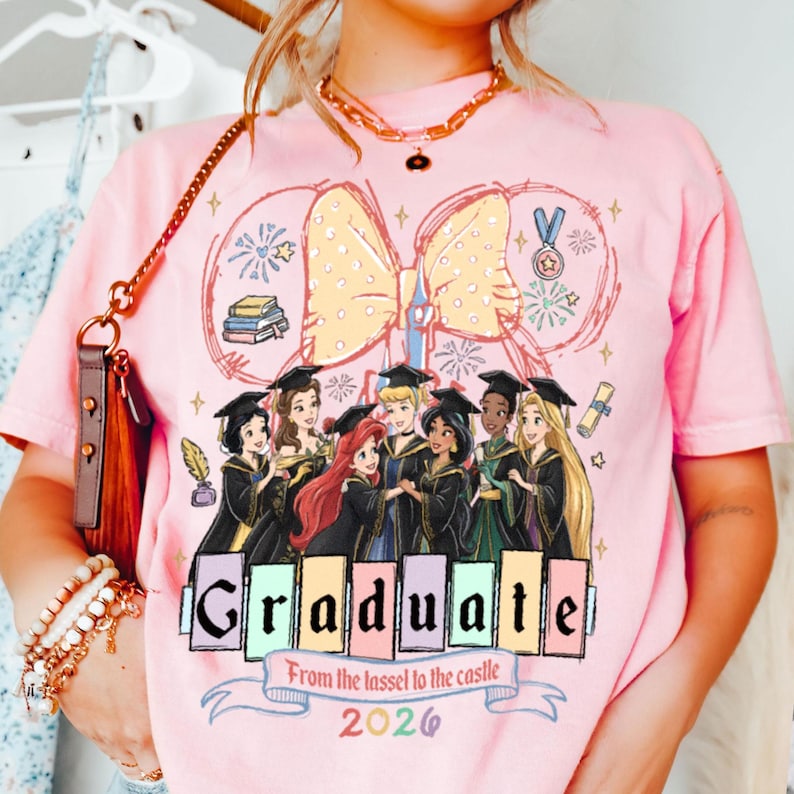 Puede incluir: Camiseta rosa con un dise&ntilde;o de graduaci&oacute;n con princesas de Disney. La palabra "Graduate" se muestra en una fuente colorida, con el texto "From the tassel to the castle 2026" debajo. Un gran lazo amarillo est&aacute; en la parte superior.
