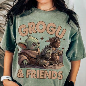 K&ouml;nnte beinhalten: Salbeigr&uuml;nes T-Shirt mit dem Text "GROGU & FRIENDS" und einer Grafik von Grogu und drei weiteren Charakteren. Die Grafik hat einen Vintage- und Used-Look. Das Shirt ist ein bequemer, l&auml;ssiger Stil.