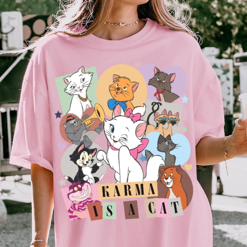 Puede incluir: Camiseta rosa con un dise&ntilde;o de dibujos animados de varios gatos. El texto "Karma is a cat" est&aacute; impreso en la camiseta.
