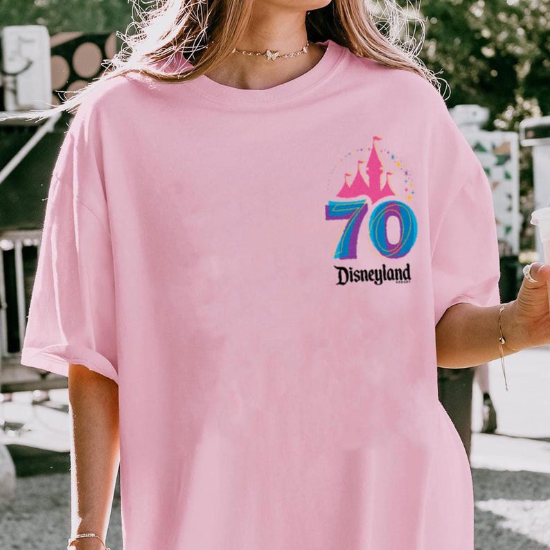 May include: A pale pink t-shirt with a colourful graphic of a castle, the number 70, and the word "Disneyland". The graphic is in shades of pink, blue, and purple. The shirt has short sleeves.