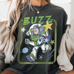 May include: A charcoal grey t-shirt featuring a graphic print of the character Buzz Lightyear in a running pose. The design includes the word BUZZ in green letters, a yellow star, a blue planet with rings, and smaller star shapes inside a green frame.